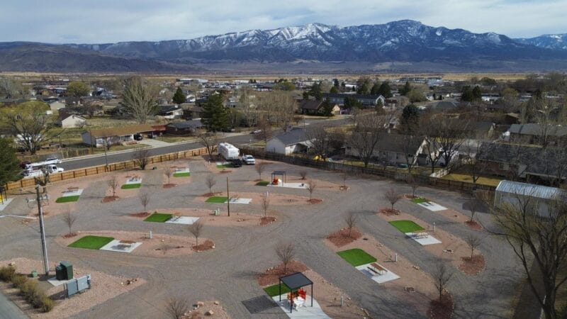 Aerial view of an RV park with multiple empty parking pads, small patches of artificial grass, trees, nearby houses, and mountains in the background.