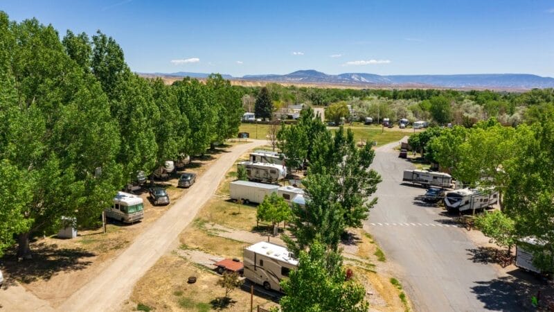Aerial view of an RV park with several motorhomes parked among green trees on a sunny day, with mountains visible in the background.