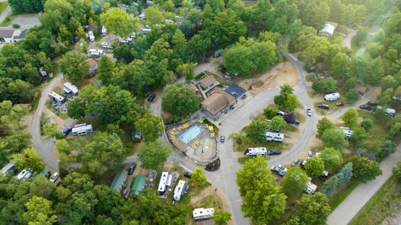 Aerial view of a campground with numerous RVs, trees, winding roads, and a central swimming pool surrounded by lounge chairs and buildings.
