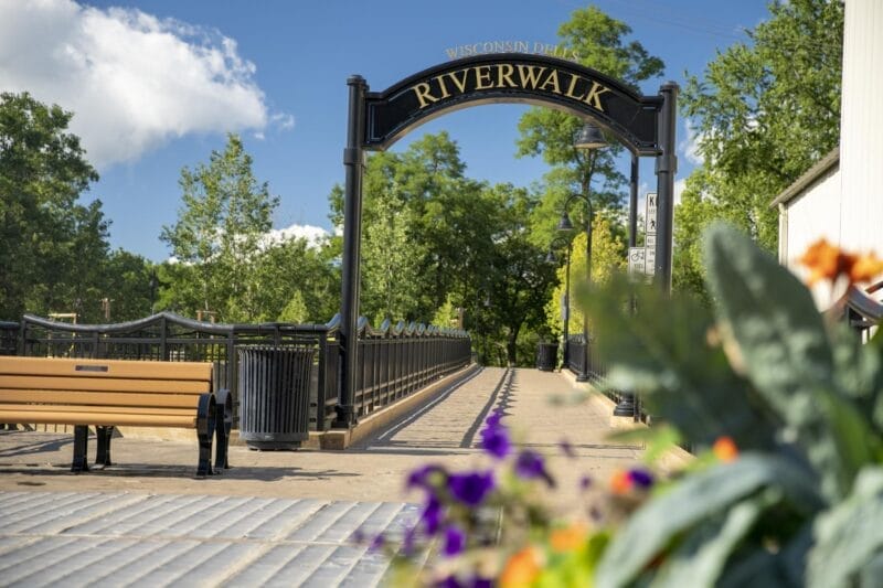 A paved pedestrian bridge with an archway sign reading "Wisconsin Dells Riverwalk," surrounded by trees and flowers, with benches along the path.