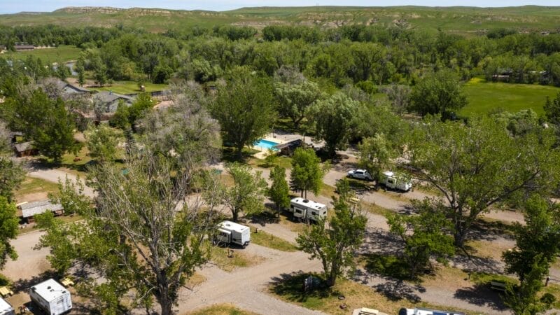 Aerial view of an RV campground with several RVs parked among trees, gravel roads, a swimming pool, and open green fields in the background.