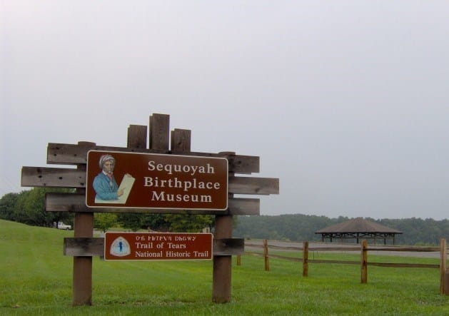 A wooden sign reads “Sequoyah Birthplace Museum” and “Trail of Tears National Historic Trail” beside a grassy field, fence, and a lakeside pavilion in the background.