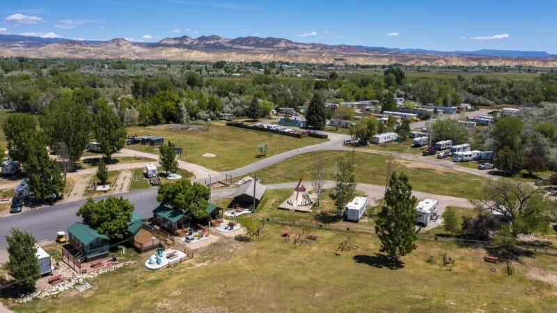 Aerial view of a campground with RVs, small cabins, trees, open grassy areas, and mountains in the background under a clear blue sky.