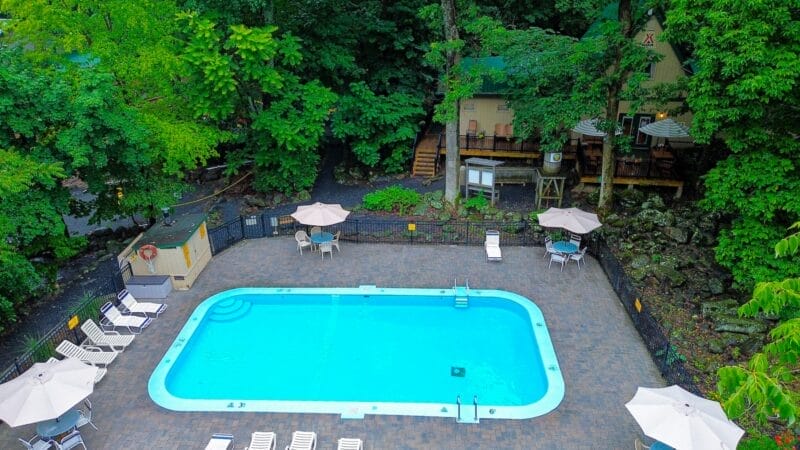 A rectangular outdoor pool with clear blue water, surrounded by lounge chairs and umbrellas, bordered by trees and a cabin in the background.