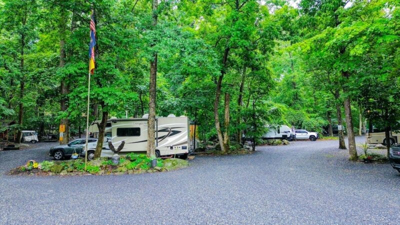 A campground with several RVs parked among tall green trees, gravel roadways, and an American flag on a pole in the foreground.