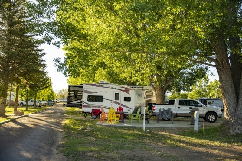 A white RV and truck parked at a shaded campsite with picnic tables, colorful chairs, and trees in the background.