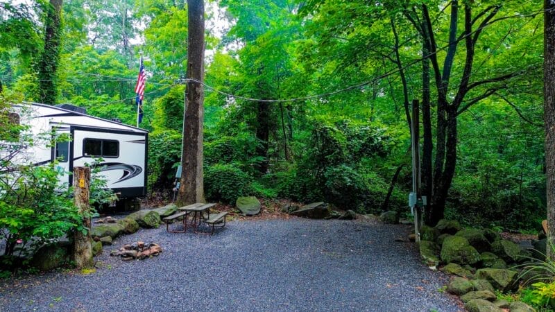 A white RV is parked at a wooded campsite with picnic tables, a fire ring, an American flag, and surrounding green trees and rocks.