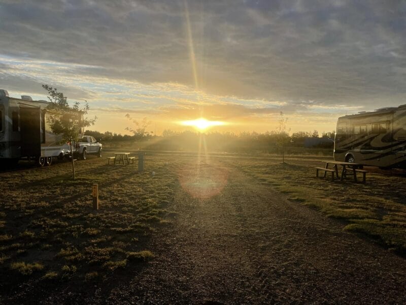 Sunset over a campground with RVs parked on either side, a gravel road in the center, and picnic tables nearby under a partly cloudy sky.