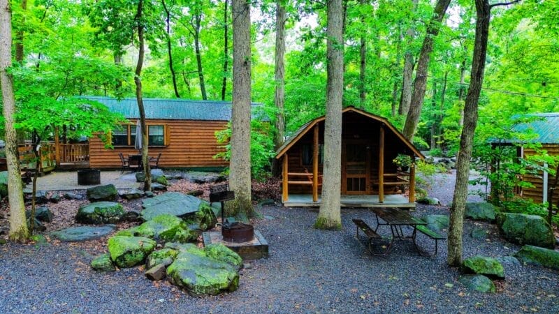 Two wooden cabins are surrounded by trees and large rocks in a forested area, with picnic tables, grills, and gravel pathways visible.