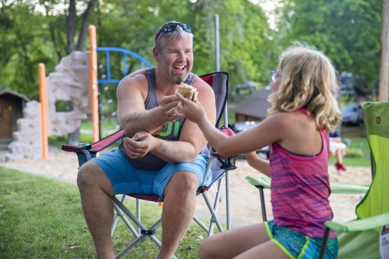 An adult and a child sit on lawn chairs outside, smiling and sharing an ice cream cone near a playground and trees in the background.