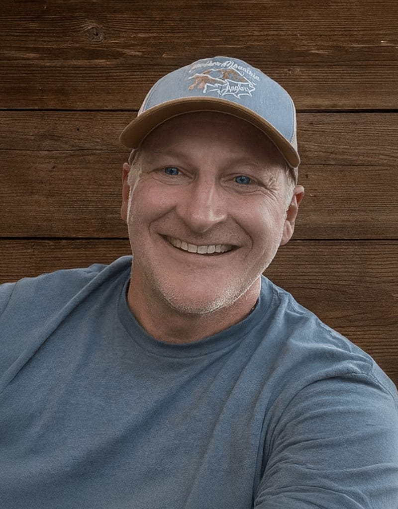 A man wearing a blue T-shirt and a blue cap smiles while sitting in front of a wooden wall.