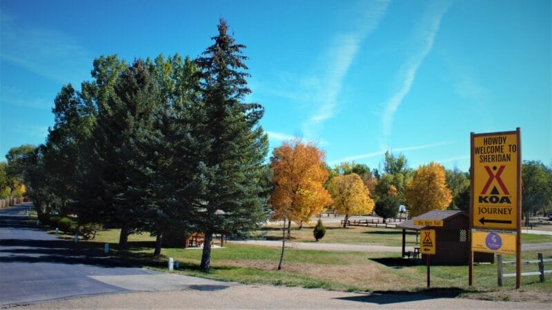 A KOA campground entrance in Sheridan, Wyoming, with a large yellow welcome sign, trees with fall foliage, and picnic tables visible in the background.