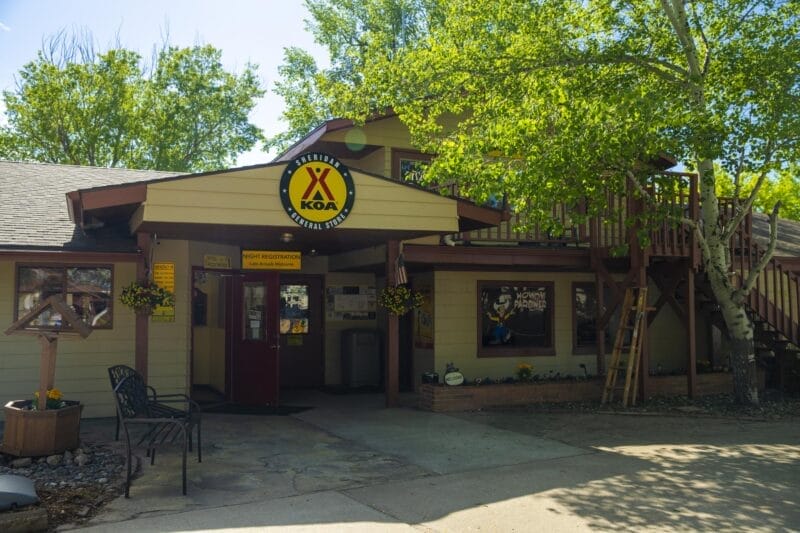A KOA campground office building with yellow signage, outdoor chairs, hanging flower baskets, and a staircase leading to an upper level, surrounded by trees.