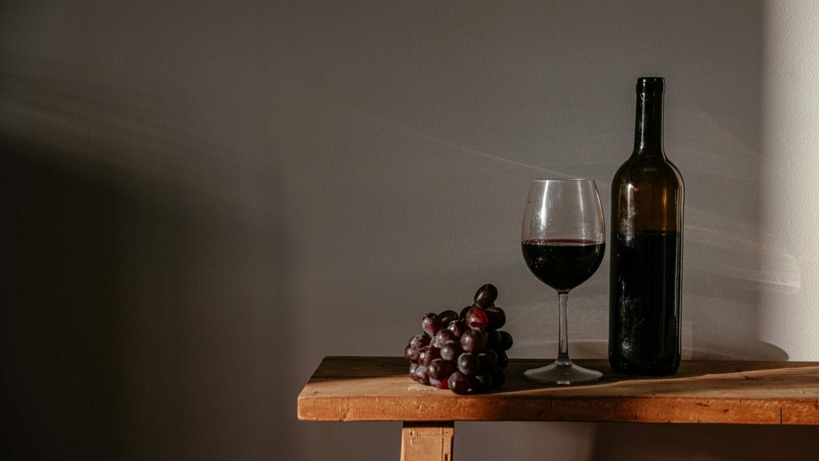 A bottle of red wine, a glass of wine, and a bunch of red grapes rest on a wooden table against a plain gray wall.