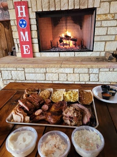 A tray of assorted barbecue meats, sides, and bread sits on a wooden table in front of a lit fireplace with a "HOME" sign on the brick wall.