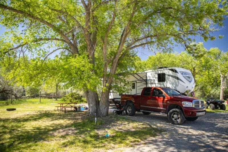 A red pickup truck is parked next to a large RV under a leafy tree at a campground, with a picnic table and grassy area nearby.