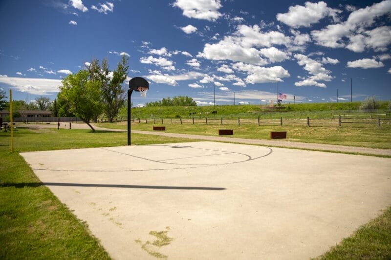 An outdoor basketball court with one hoop is set in a grassy park area under a blue sky with scattered clouds; a flag and fence are visible in the background.