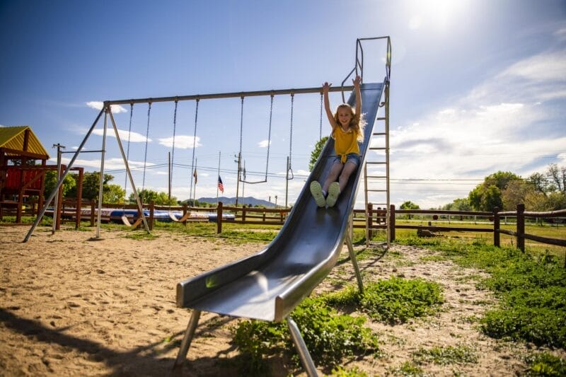 A young girl in a yellow shirt slides down a metal playground slide on a sunny day, with empty swings and a sand-covered ground in the background.