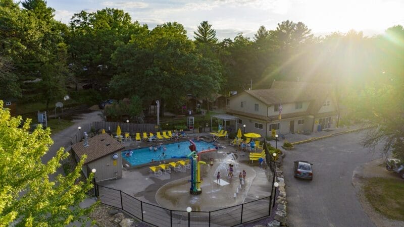 Aerial view of an outdoor pool and splash pad area with people, surrounded by trees, yellow chairs, and nearby buildings on a sunny day.