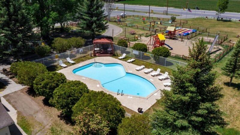 A fenced outdoor swimming pool with lounge chairs next to a playground area, surrounded by trees and located near a road.