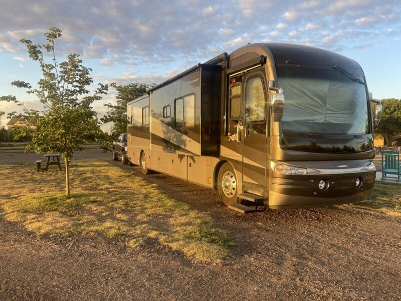 A large brown and beige RV is parked on a gravel lot with a small tree, picnic table, and grassy area nearby under a partly cloudy sky.