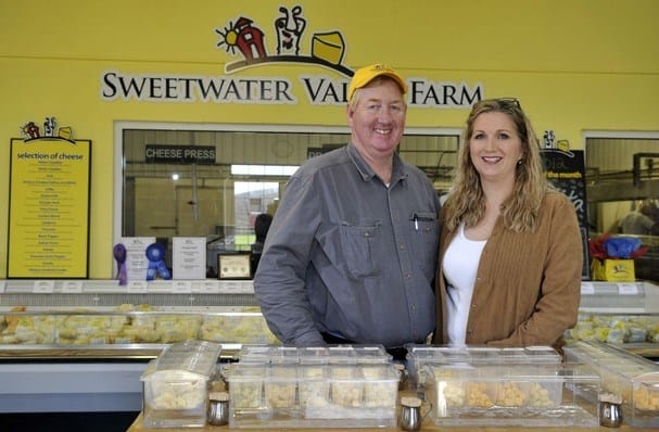 Two people stand behind a counter with cheese samples at Sweetwater Valley Farm, with a cheese display and awards visible in the background.