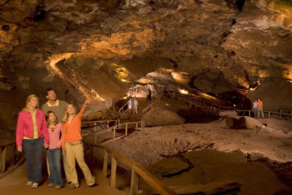 A family stands on a walkway inside a large, well-lit cave with other visitors exploring in the background.