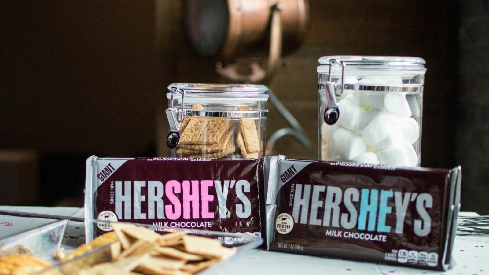 Two large Hershey's milk chocolate bars are on a table in front of jars containing graham crackers and marshmallows.