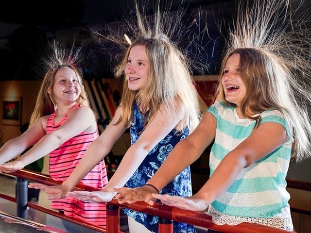 Three girls stand with their hands on a metal rail, hair standing on end due to static electricity, all smiling and laughing indoors.