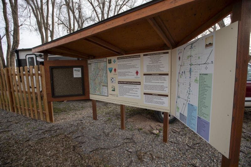 Covered outdoor information board displaying maps, notices, and park guidelines, located on gravel near a wooden fence and trees.