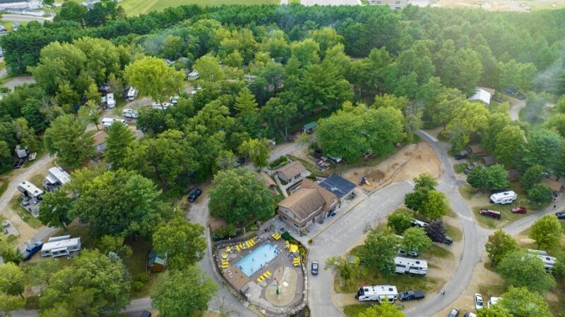 Aerial view of a campground with RVs, a central swimming pool, and several buildings surrounded by dense trees and winding roads.