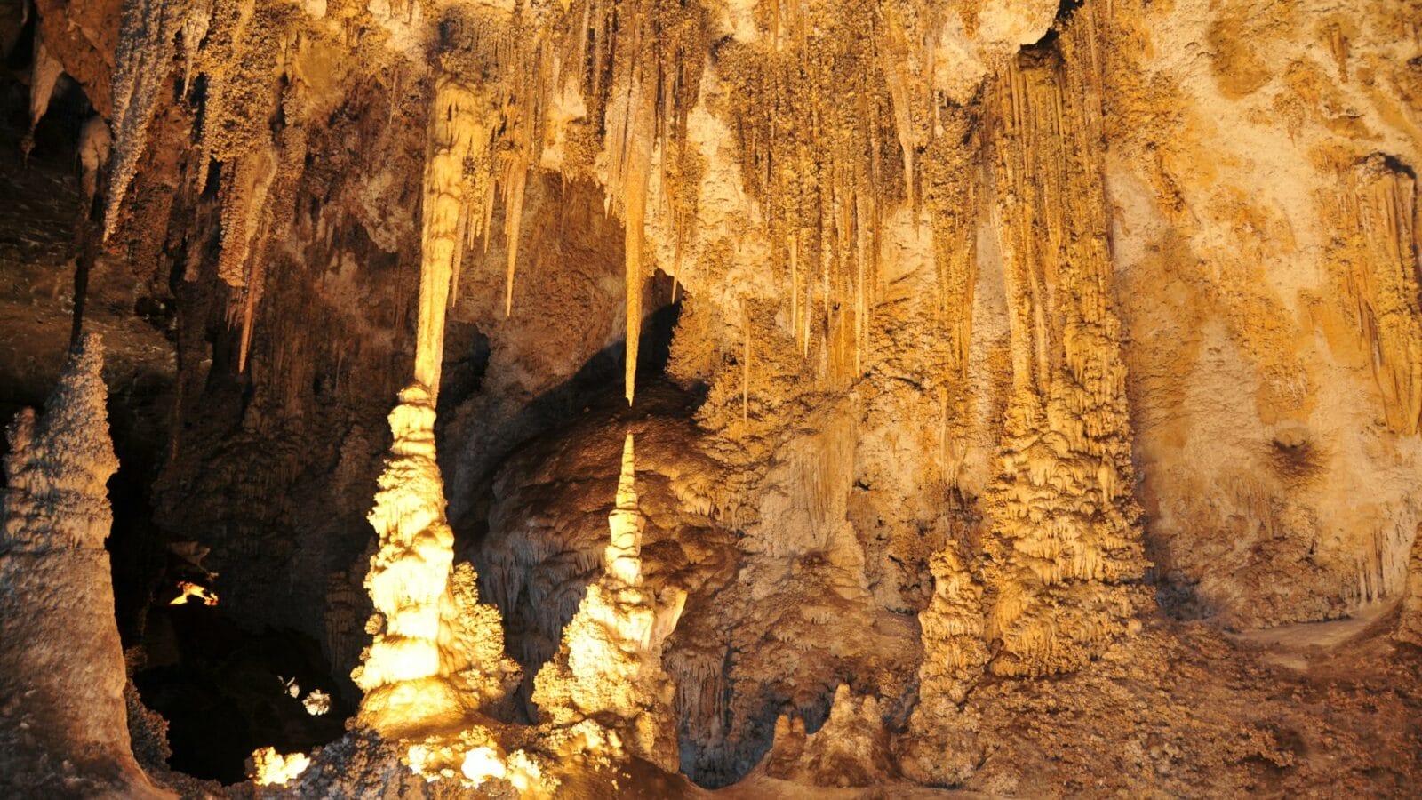 A cave interior with limestone stalactites hanging from the ceiling and stalagmites rising from the floor, illuminated by warm artificial lighting.