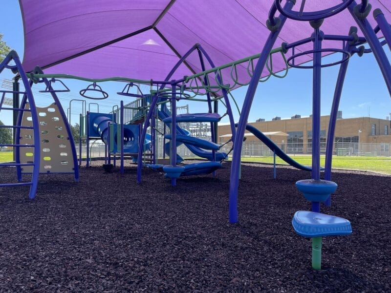 A modern playground with slides, climbing structures, and monkey bars is shaded by a large purple canopy. The ground is covered with dark mulch. A school building is visible in the background.