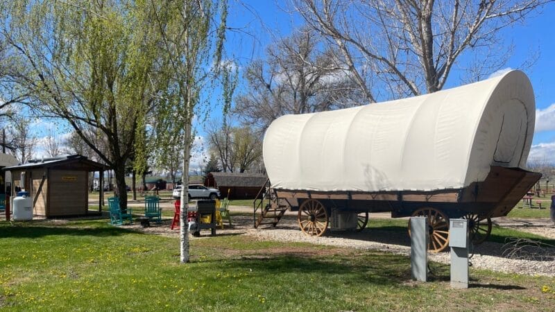 A covered wagon on display in a grassy park area with picnic tables, trees, a small wooden building, and blue sky.