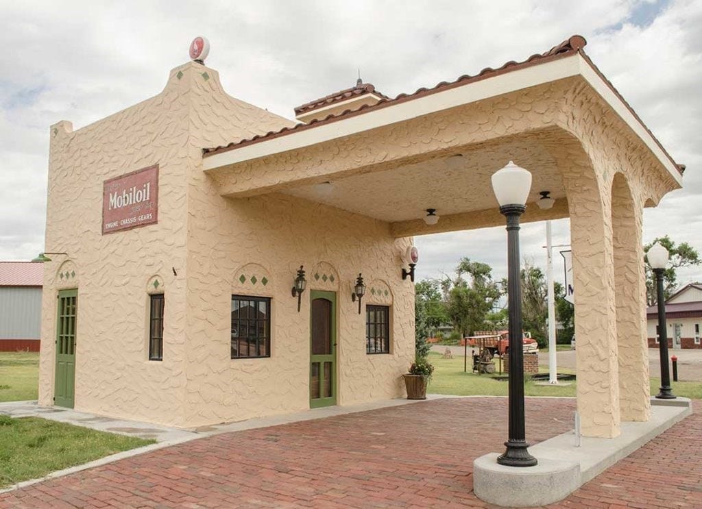 A historic tan stucco building with a covered drive-through area, green-trimmed doors, and a Mobil oil sign above the entrance.