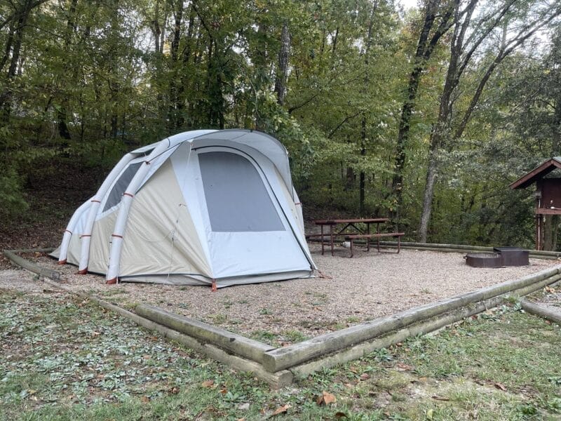 A white tent is set up on a gravel campsite with a picnic table and fire ring, surrounded by trees and bordered by wooden beams.