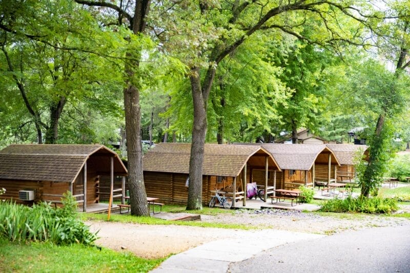 Several wooden camping cabins with picnic tables are lined up under tall trees in a green, wooded area along a paved path.