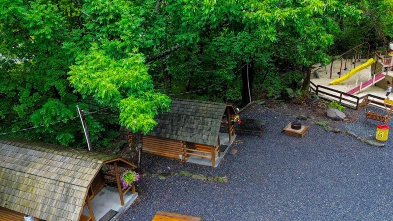 Aerial view of two small wooden cabins near a gravel area, surrounded by trees, with a playground and yellow slide in the background.