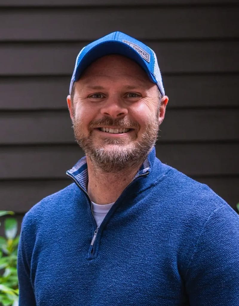 A man wearing a blue cap and zip-up sweater smiles in front of a dark paneled background.