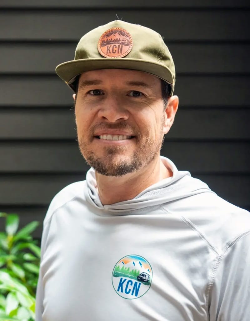 A man wearing a green cap and a gray sweatshirt with a "KCN" logo stands in front of a dark wall, smiling slightly.