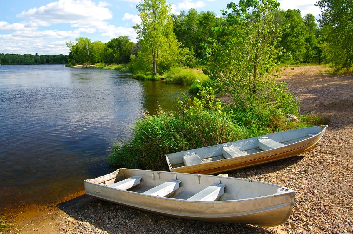 Two empty aluminum rowboats rest on a pebble shore beside a calm river, surrounded by green trees under a partly cloudy sky.