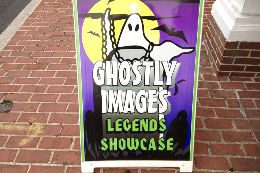 A signboard with a cartoon ghost, bats, and full moon reads "Ghostly Images Legends Showcase" on a brick sidewalk.