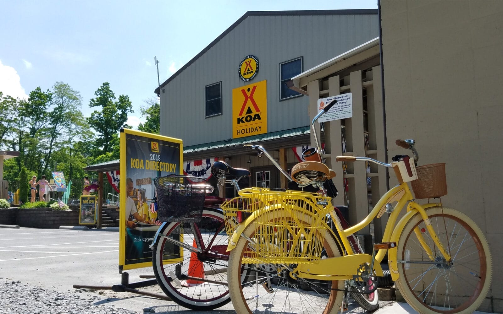 Two yellow bicycles are parked outside Elizabethtown / Hersey KOA Holiday campground building on a sunny day, with trees in the background.
