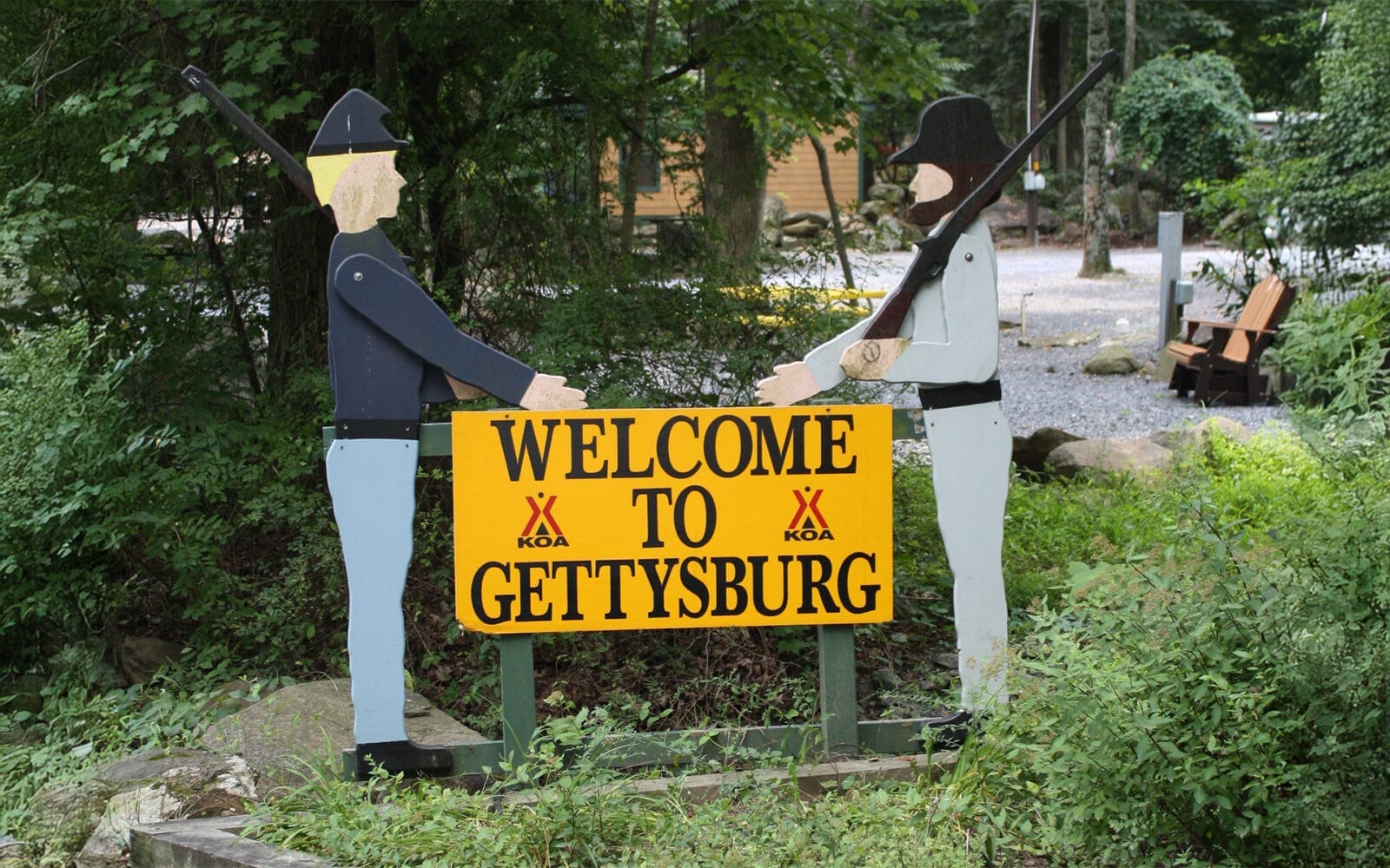 A wooden sign reading "Welcome to Gettysburg KOA" stands between two soldier cutouts, one in blue and one in gray, with trees and a campground in the background.