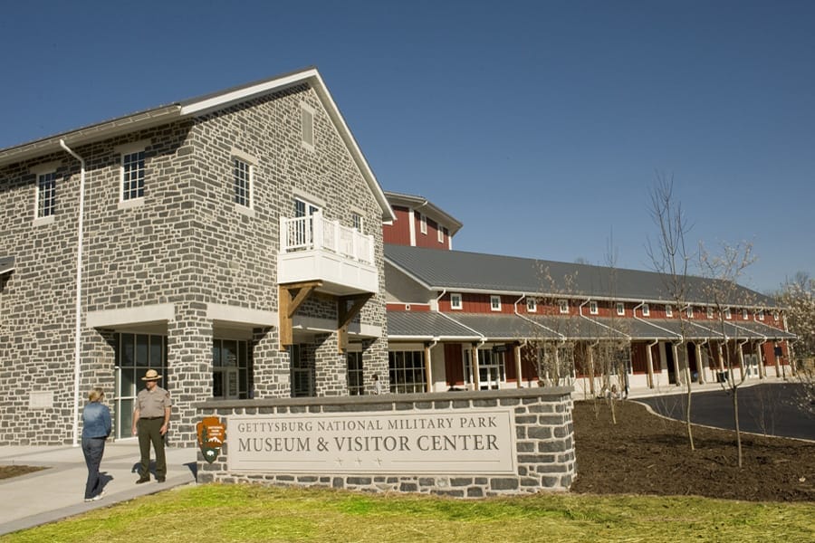 Three people walk outside the Gettysburg National Military Park Museum and Visitor Center, a stone and red building under a clear blue sky.