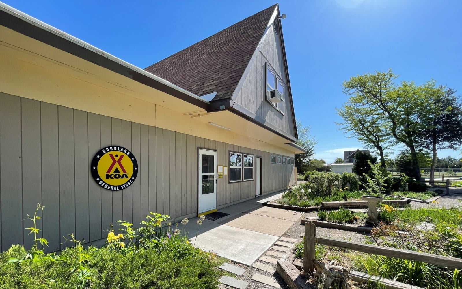 In Goodland KOA, an a-frame building with a KOA sign on the side is set against a clear blue sky. The foreground features a garden with various plants and a wooden railing.
