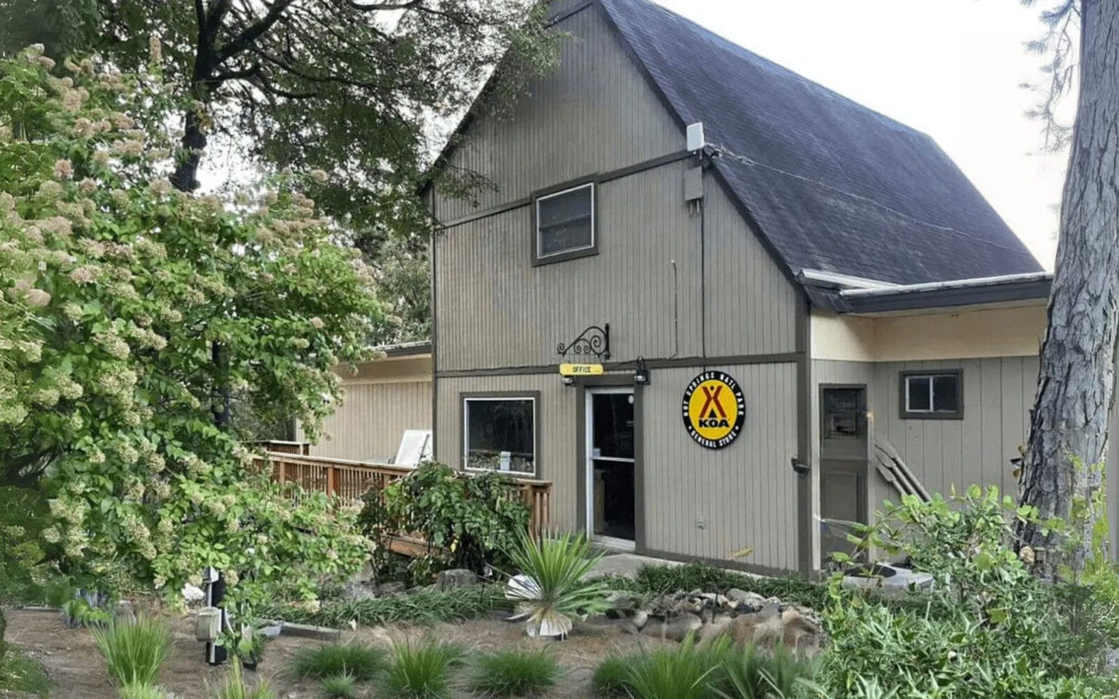At Hot Springs National Park KOA, a two-story wooden building with a pitched roof is surrounded by trees and plants. A yellow KOA sign is displayed on the wall. A wooden railing leads to the entrance.