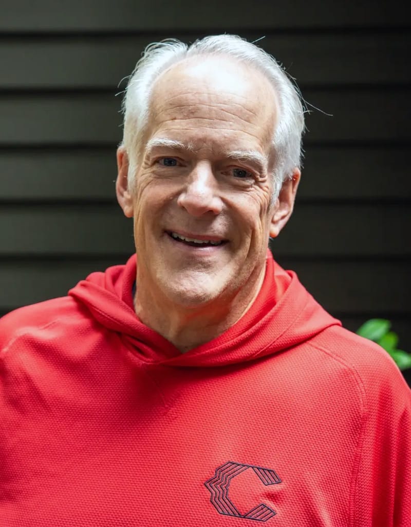 A man with white hair smiling, wearing a red hoodie with a logo on it, standing against a dark background.
