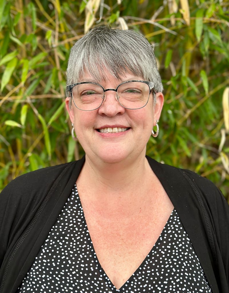 Julie has short gray hair, glasses, and hoop earrings. She is wearing a black and white polka dot top and black cardigan.