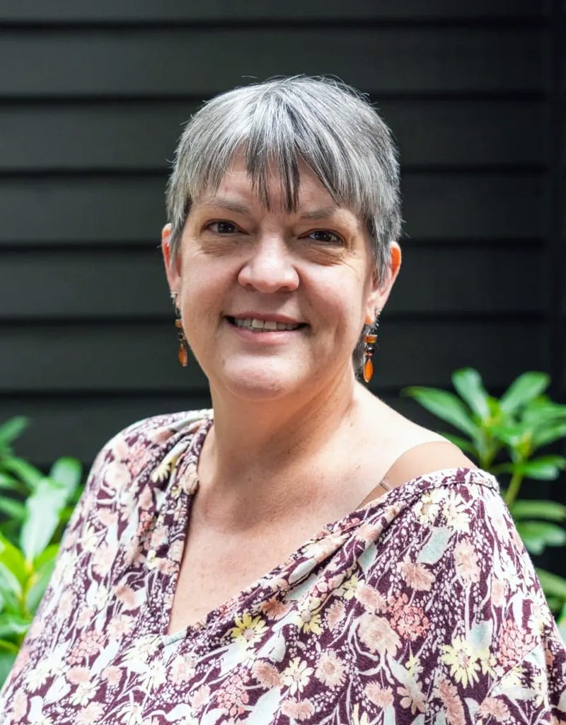 A woman woman with short gray hair and floral top stands in front of dark siding and green foliage.